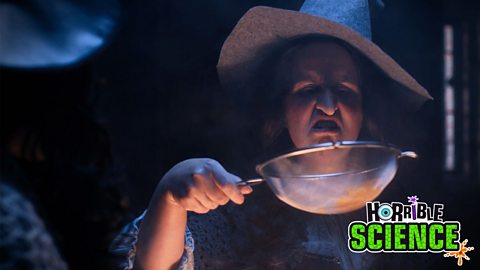 A witch in a grey hat is looking at a large sieve. 
