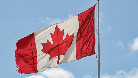 a landscape image. The Canadian flag is flying from a pole. It is split into thirds, with two bright red vertical blocks on either side and white in the middle. On the white patch is a red maple leaf.