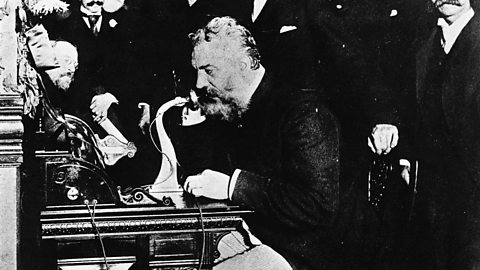 A black and white photograph of Alexander Graham Bell, an older white man, sat at a desk with an early version of the telephone as makes a long-distance call. He's surrounded by other men watching.