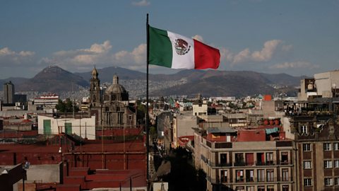 A landscape image looks out onto Mexico City, with rooftops visible against a grey-blue sky. In the foreground, the Mexican flag flies on a pole. It's a vertical tricolour with fields of green, white and red from left to right. In the centre of the white stripe is the Mexican Coat of Arms - a golden eagle on top of a pear cactus, devouring a serpent.