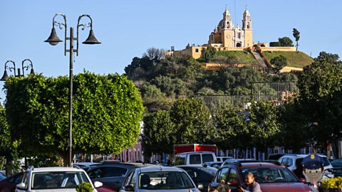 A landscape image shows the Cholula pyramid in the present day. In the foreground, cars travel along a busy road. Behind them are green trees, and in the background is the large mound of the pyramid with a medieval church on top of it.