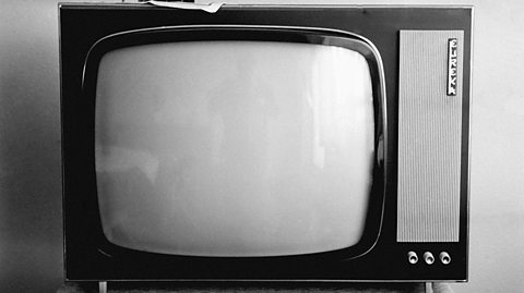 A black and white photograph of an old-fashioned television set. It's shaped like an oblong box, with a screen and a side panel that has buttons and knobs on it. 