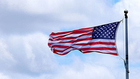 A landscape image of the American flag flying from a pole against a cloudy grey sky. The flag has 13 alternating red and blue horizontal stripes. In the top right-hand corner is a dark blue square dotted with 50 white stars.