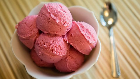 The image shows a white bowl of ice cream on a table with a silver spoon to the right. The bowl contains six coops of pink gelato.