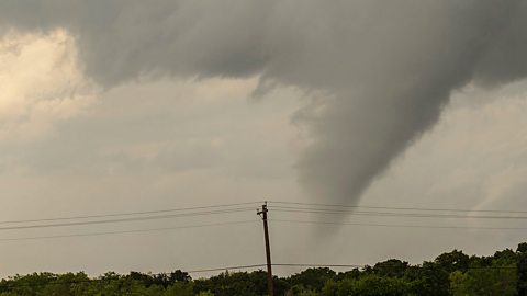 A landscape image shows the funnel of a tornado spinning up from ground level high into the sky. The tornado is dark grey against a slightly lighter grey sky.