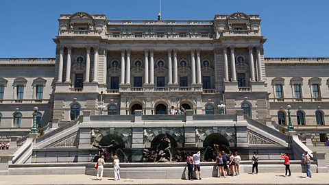 An image of the exterior of the Library of Congress. It's a large Gilded Age building with a facade of white granite, a large split staircase and a fountain. A few people are gathered at the bottom of the image.