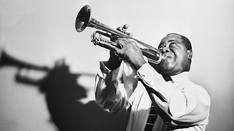 A black and white publicity image of the African American jazz musician Louis Armstrong. He is playing a trumpet and wearing a light-coloured shirt.