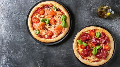 An image shows two pizzas on separate plates on a dark grey table. The picture has been taken from above.