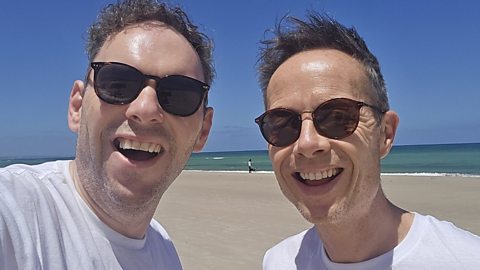 Two men on a beach, each wearing sunglasses and smiling to camera.