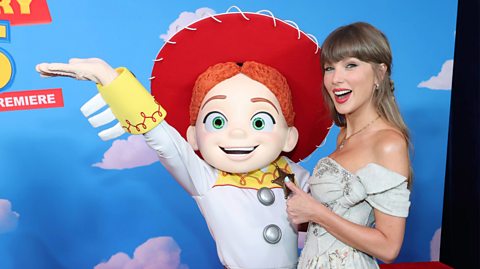 Against a blue sky background with clouds, a lifesize costume of Toy Story's Jess, with a red cowgirl hat, middle parted red hair and a white buttoned shirt with yellow cuffs, stands next to a smiling Taylor Swift. Taylor has blonde hair with a fringe and wears a white strapless dress and smiles with her thumb up for the camera
