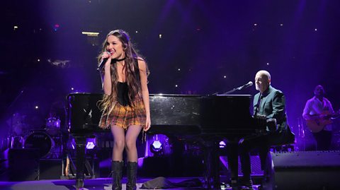 Olivia Rodrigo and Billy Joel perform "Deja Vu" and "Uptown Girl" onstage at Madison Square Garden. On stage, Olivia sings into a mic wearing a short checked yellow skirt, a black top, black choker necklace and black boots. She has long brown hair. Behind her is Billy Joel, who wears a suit and sits seated behind a piano playing the keys