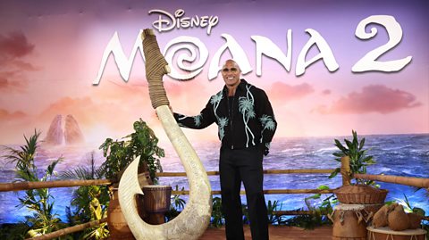 Dwayne 'The Rock' Johnson attends the UK Premiere of Moana 2. He wears dark sunglasses, black trousers and a black shirt with blue detail. He stands in front of a poster advertising the film posing next to a large fish hook associated with the character he voices in the film