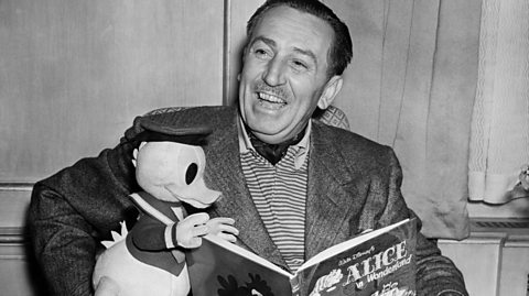 A black and white photo of Walt Disney smiling with a Donald Duck toy while reading Disney's Alice in Wonderland book. He wears a striped polo shirt and a blazer