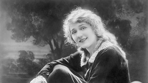 A black and white photograph of Mary Pickford, who is sat with her legs drawn up to her chest and her arms around them. She is gazing at the camera with a faint smile. She has long light-coloured hair.