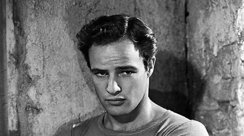 A black and white photograph of actor Marlon Brando. It's a head and shoulders photo. He's looking moodily off to the right of the camera with his lips pursed.