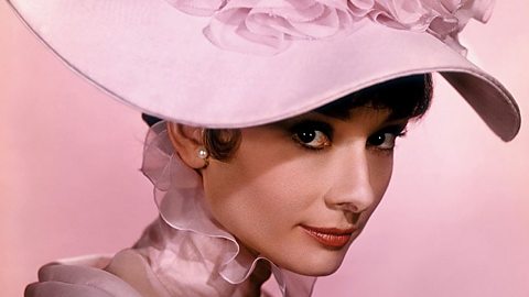 A colour photograph of actress Audrey Hepburn, wearing a fashionable wide-brimmed light pink hat. It covers part of her face and she's looking at the camera from under the brim with a small, coquettish smile.
