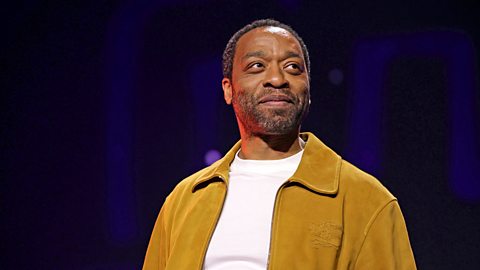 A landscape image of actor Chiwetel Ejiofor, who is wearing a tan brown jacket over a white T-shirt. He's looking off to the right and smiling.