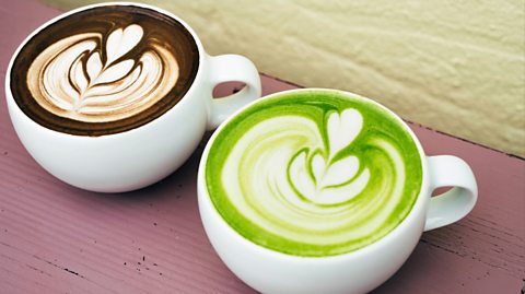 On a wooden table is two white mugs, one with a specialty hojicha latte which is brown and the other a green matcha latte. They both have coffee art on top of them, with the milk added forming a leaf pattern on top