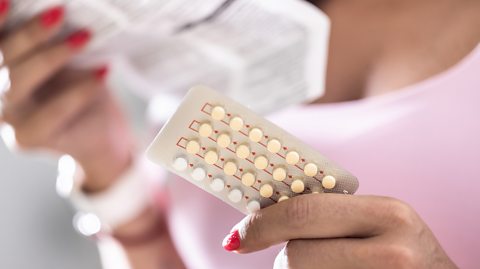Hormone contraceptive tablets unopened in a blister pack, held by a woman wearing a pink top blurred in the background
