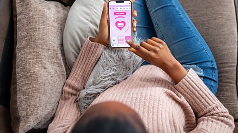 A woman wearing a pink jumper and blue jeans lies on a couch on her phone. She is using an app to help keep track of their periods which has a pink screen
