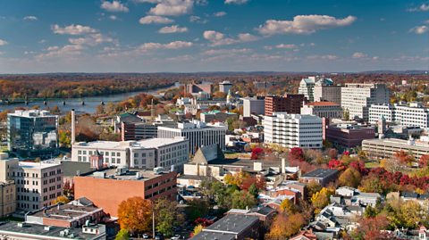 An aerial view of Downtown Trenton, New Jersey on a sunny day with blue sky, buildings and green and red leaved trees in the distance