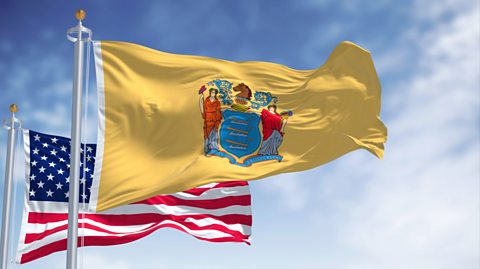 The New Jersey state flag and the USA flag. The state flag is buff, a yellow-ish tan colour, and dark blue, known as ‘Jersey blue.’ t features two female figures either side of a shield which has three ploughs at the centre, a horse’s head above a knight’s helmet and a blue ribbon displaying the state motto of ‘Liberty and Prosperity.’ The USA flag behind has white stars on a blue background in the left hand corner, with the remainder of the flag being red and white striped