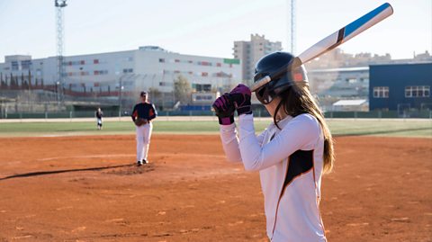 A woman wearing all white, gloves and a helmet holds a bat ready for the pitcher to throw a baseball for her to bat. They are on a sandy baseball track