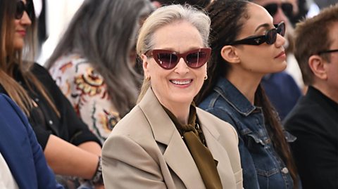 Actress Meryl Streep smiles for the camera wearing her white hair in a ponytail, red sunglasses, a beige blazer and a khaki tie front blouse
