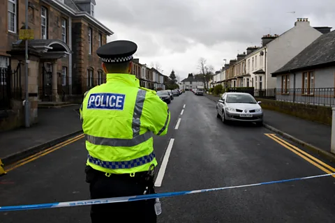 Getty Images A police officer in a high-visibility jacket standing behind blue and white police tape closing off a residential street in Glasgow (Credit: Getty Images)