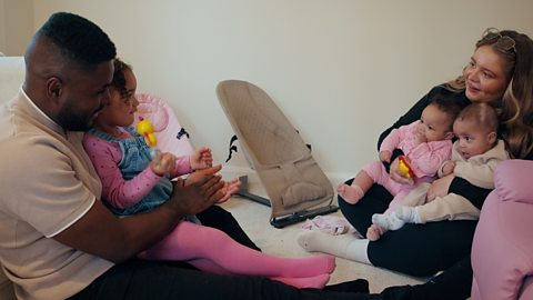 A father on left supports daughter by holding her on lap; he's facing towards his partner to the right, the mum, who is supporting their other two daughters.