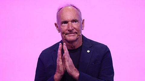 An image of Tim Berners-Lee. He's a white man in his early seventies. He's stood against a light pink background wearing a dark shirt and is clapping his hands together. He's speaking at an event.