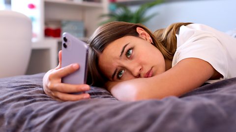 A landscape image that shows a teenage girl lying on her bed with her head resting on her arm. She's wearing a fed up expression while staring at her phone, which she's holding up in front of her face.