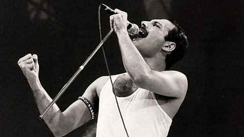 Freddie Mercury, lead singer of Queen, on stage wearing a white vest and arm band accessory. He has dark hair and a dark moustache and sings into a mic and stand which he lifts into the air
