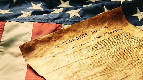 On an American flag, which has white stars on a blue background and red and white stripes, lies an old document stained and crumpled which appears to be the Declaration of Independence