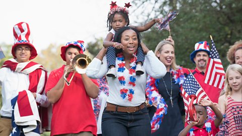 A group of adults and children smile as they walk during a Fourth of July celebration parade. In various ways, they all wear red, white and blue and some are waving USA flags in their hands