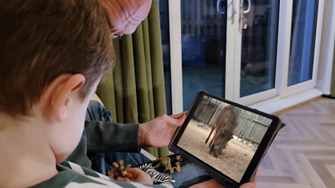 Child looking at a video tablet, that has a wart-hog on the screen. His dad is holding the screen with his left hand.