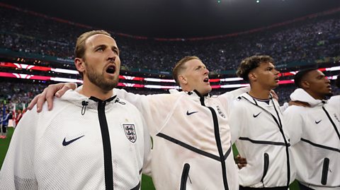 The England men's football team sing the national anthem during a World Cup 2026 game. The team all wear white tracksuit jackets and sing with their hands around one another on the football pitch
