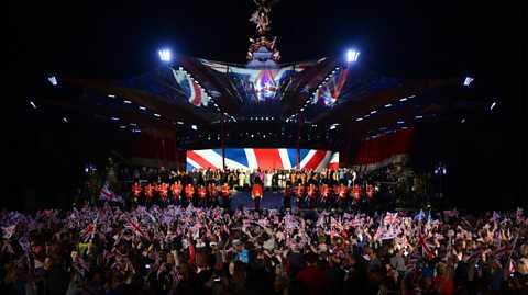 Queen Elizabeth II, Prince Charles, Prince of Wales, Camilla, Duchess of Cornwall, and performers stand on stage as God Save The Queen is sung at the end of the Queen's Diamond Jubilee Concert, 2012. At nighttime, there is a large crowd watching and the screen on stage is projecting the Union Jack flag