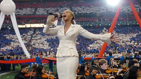 Beyonce sings the National Anthem to kick off Super Bowl XXXVIII, 2004. She has her blond hair in a ponytail and sings into a mic wearing a white suit-style jacket and matching pencil skirt. On the American football field is violinists behind her and red, white and blue decorations to symbolise the American flag.