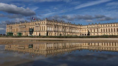 A landscape image in colour shows the exterior of the Chateau de Versailles. It is built in the symmetrical French classical and Baroque styles. It features a large golden entry gate, rows of arched windows and stone facades with columns and statues. Its image is reflected in a large body of water in front of it. 