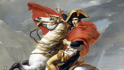 A colour portrait of Napoleon Bonaparte. He is dressed in military clothing and is sat upon a large white horse, which is rearing up with its front legs in the air. 