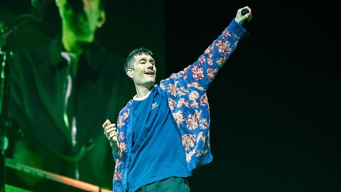 An image in colour shows Dan Smith, the lead singer in the band Bastille, performing on stage. He's looking off to the right towards his arm which is raised in the air. 