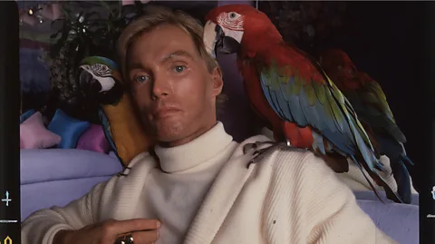 HBO Frederick Von Mierers in a white turtleneck and coat, with two colourful parrots on his shoulders (Credit: HBO)