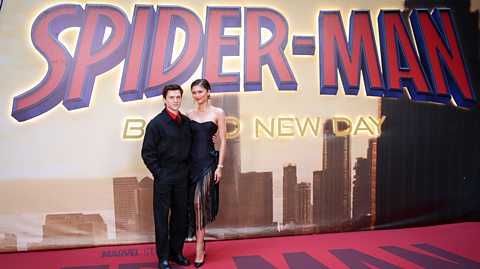 Tom Holland and Zendaya at the premiere of Spider-Man: Brand New Day, 2026. On the red carpet, Tom wears a black suit with a red shirt and Zendaya wears a strapless black dress. Behind them is a poster advertising the film which shows the text of the film over the New York City skyline