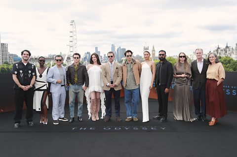 Cast members of Sir Christopher Nolan's The Odyssey pose in a straight line for a photo. They stand on a black carpet with 'The Odyssey' printed on it in white text and behind them is the London skyline, including the London Eye.