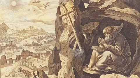 Alamy A detail of an artwork depicting medieval priest John Cassian sitting at the entrance of a cave on a clifftop overlooking a view of mountains, buildings, sun and sky. There is a book in his lap (Credit: Alamy)