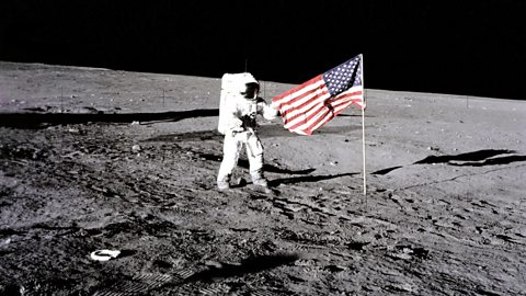 A grainy image from the second American moon landing in 1969 shows an astronaut dressed in a white space suit unfurling the American flag, which has been planted on the surface of the moon. The ground is grey and craggy and the sky is completely black. 
