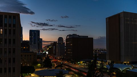 A landscape image shows Phoenix, Arizona at night. The sky is a greyish-blue, giving way to a lighter pinkish-orange. In the foreground, tall buildings are silhouetted against the sky, and beneath them we can see lights from windows and traffic. 