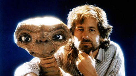 An image shows director Steven Spielberg holding the head of the puppet used to portray E.T. One hand is holding the puppet and the other stroking his chin thoughtfully. He's gazing off slightly to the left of the camera. 