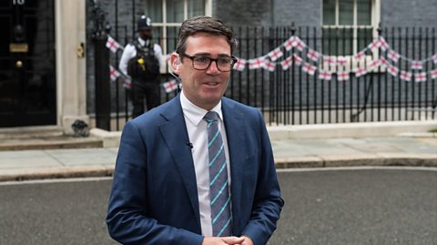 Prime Minister Andy Burnham outside Downing Street being interviewed outside 10 Downing Street. Behind him is the number 10 black door, a police officer and the railings with England flag bunting. He has dark hair, glasses and wears a navy suit blazer, a white shirt and grey and blue striped tie.